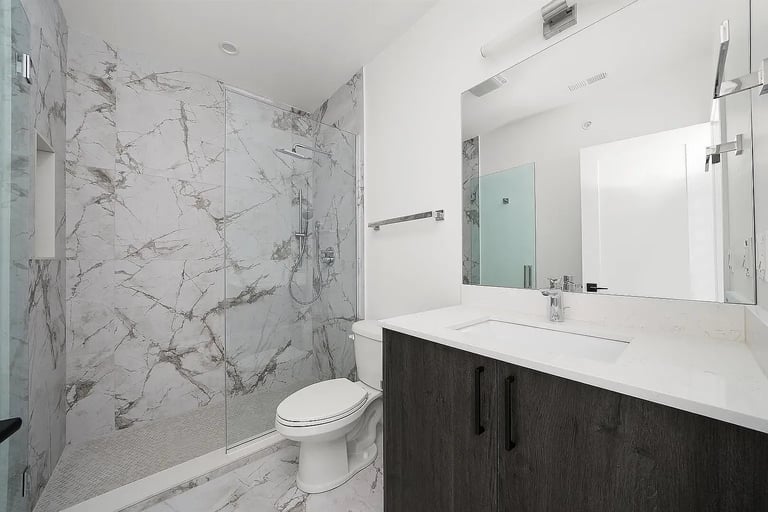 Modern bathroom with marble-tiled shower enclosure, white toilet, black vanity with white countertop, and large mirror